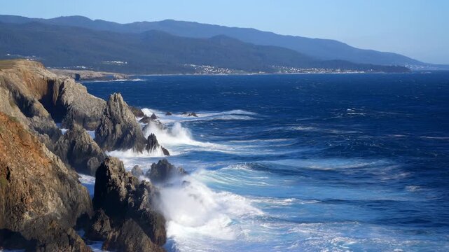 Powerful ocean waves crash against jagged rocky cliffs, throwing white spray across deep blue water while distant mountain ridges and shoreline stretch beneath clear daylight