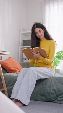 Young Latino female college student sitting on bed and reading a book. 