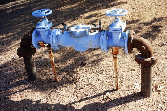 Blue painted water backflow preventer assembly with two handwheel shutoff valves and rusty plumbing pipes on ground outdoors ; backlit