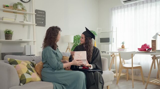 Senior Latino mother give gift box to young daughter in graduation gowns.