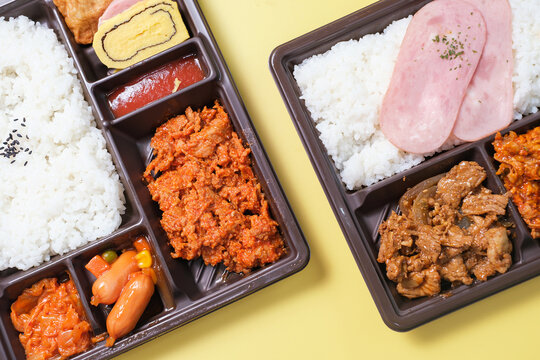 Korean lunch box with rice, meat, and kimchi in plastic packaging on yellow background close-up, top view.