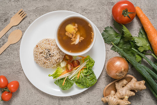 Ginger chicken soup served in a bowl, accompanied by red rice and a side salad arranged on a plate, complete with fresh ingredients used as props.