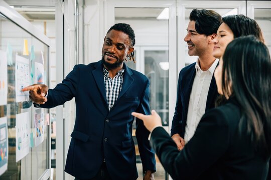 A diverse group of professionals in a business office, focusing on discussions around a board. The black businessman emphasizes teamwork, leadership, and effective planning. team work