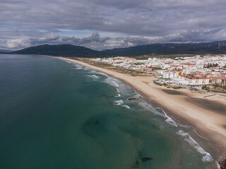 Aerial view of a long sandy beach with turquoise ocean waves meeting a coastal town beneath dramatic cloudy skies and distant mountains in Tarifa, Spain © Felix Morion