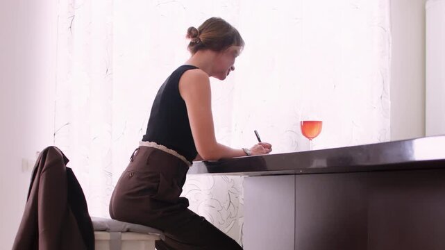 Professional journaling. Daily planning. Thoughtful stylish woman writing notes at home table enjoying orange drink in glass interior.