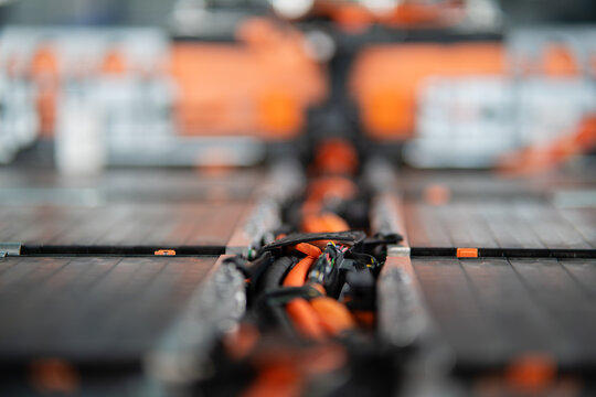 An electric vehicle battery pack reveals orange high voltage wiring and connectors between its exposed modules in shallow focus.