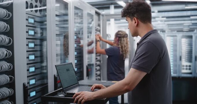 Male IT Engineer Reviews Server Telemetry on Laptop While Female Colleague Works Behind Him and Discusses Infrastructure Status, Showing Coordinated Diagnostics and Maintenance in Modern Data Center.