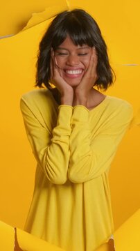 Joyful young Asian woman looks genuinely happy, resting her hands on her cheeks with a beaming smile, surrounded by torn yellow paper. Woman radiates positivity and sweet, charming expression.