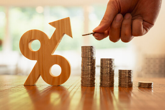 Interest rate increase and inflation concept, hand putting coin on growing stack of money beside wooden percentage arrow symbol on table, representing financial loan, mortgage, and investment.
