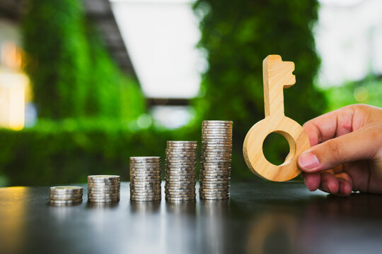 Real estate investment and financial success concept, hand holding wooden key beside ascending stacks of coins on table, symbolizing home loan, property purchase, and mortgage growth.