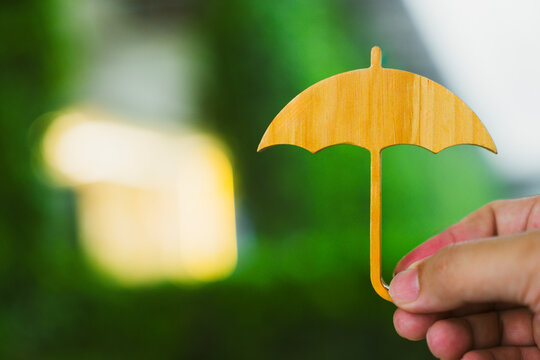 Insurance protection and security concept, hand holding small wooden umbrella cutout on blurred green nature bokeh background, symbolizing safety coverage, health safeguard, and care.