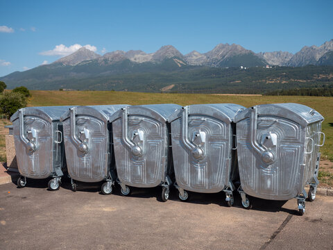 Row of large metal garbage containers on wheels in a parking lot with mountains in the background, combining municipal waste management infrastructure with a scenic rural landscape.
