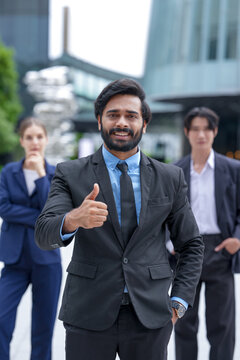 Three young business professionals wearing conference badges fold their arms confidently for a team photograph on the open terrace of a modern corporate campus in the city centre
