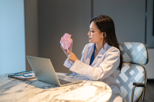 Asian female cardiology doctor holding a pink heart model for online telehealth on laptop, Medical expert explaining heart attack disease, patient education and digital healthcare consulting concepts