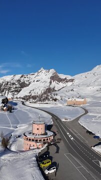 The road over the Simplon Pass runs past a round stone tower and on to the old hospice, Switzerland, cars parked on cleared tarmac between deep winter snowfields. Aerial drone wide shot