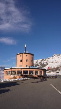 The round pink pass building at the Simplon Pass, Switzerland, stands under a deep blue winter sky with a Swiss flag and snow covered rock peaks behind it. Static wide shot