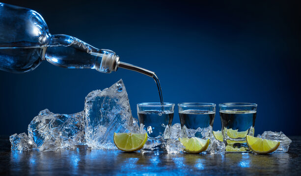 Shot glasses of vodka with lime slices and ice on a dark blue background.