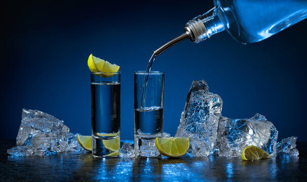Shot glasses of vodka with lime slices and ice on a dark blue background.