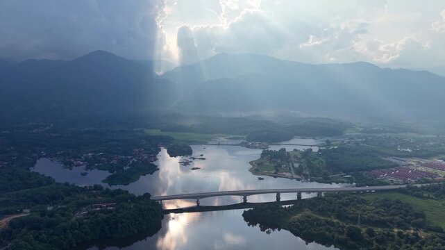 Sunlight pierces through clouds above the river. Wide bridge spans the water, connecting villages at Tasik Raban