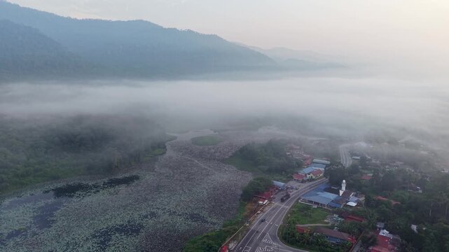 A misty morning landscape showing a winding road passing through a green valley with forested hills and scattered houses at Tasik Raban