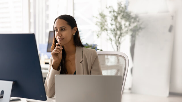 Focused female office employee reviewing information on computer screen, thinking through decision or solution, analyzing work results or digital data, planning business strategy or preparing report