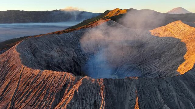 Golden sunrise light illuminates steaming Mount Bromo crater rim with dramatic textures and distant volcanic peaks.