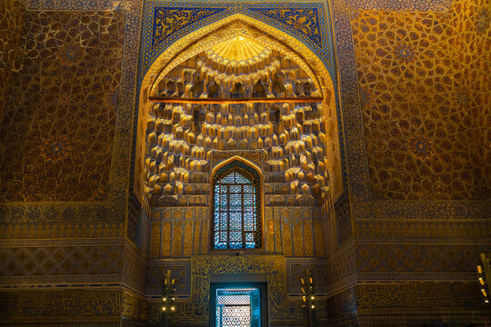 Interior architectural detail from the Amir Temur Mausoleum in Samarkand, showing a recessed arched niche covered with three-dimensional golden muqarnas, blue-and-gold geometric tilework, floral patte
