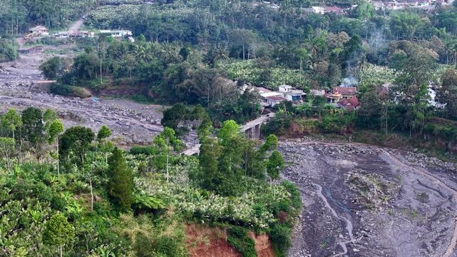 Aerial drone view of a dark, dried volcanic lava flow path from Mount Semeru, showing solidified volcanic rock and sediment cutting through the landscape.