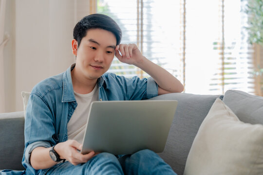 Young Asian man relaxing on comfortable couch, using laptop at home, happy male chatting with friends in social network, spending weekend, watching movie, shopping online