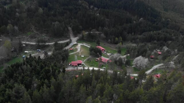 Drone footage captures a small mountain village with red-roofed buildings nestled in dense forest in Livaditis, Greece. The camera moves laterally across the scene revealing winding mountain roads.