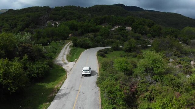 A white car travels along a winding paved road through a mountainous region with dense green vegetation and scattered trees. The shot captures the rural landscape of Livaditis village in Eastern.