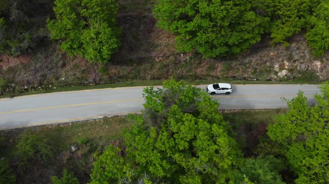 A white car travels along a curving mountain road surrounded by dense green forest in Livaditis village, Eastern Macedonia and Thrace. Overhead drone tracking shot follows the vehicle smoothly.