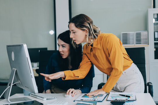 Two businesswomen discussing work using a laptop in a modern office lounge area, showing teamwork and communication
