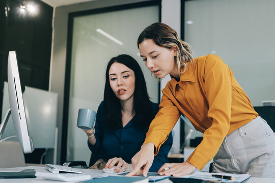Two businesswomen discussing work using a laptop in a modern office lounge area, showing teamwork and communication