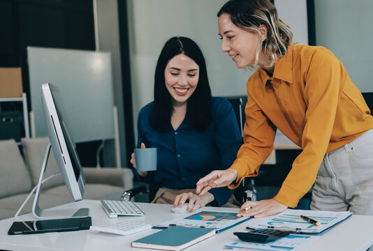 Two businesswomen discussing work using a laptop in a modern office lounge area, showing teamwork and communication