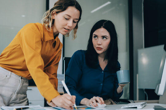 Two businesswomen discussing work using a laptop in a modern office lounge area, showing teamwork and communication