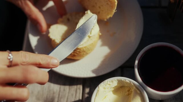 Hands spreading butter on a scone with jam and cream nearby