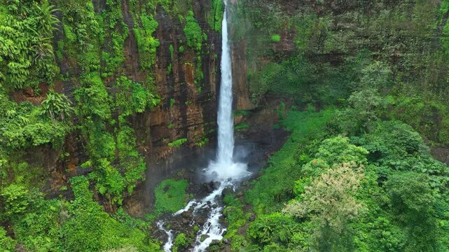 Tall Cascade With Lush Greenery And Reddish Cliff Walls At Kapas Biru Waterfall In East Java, Indonesia. Aerial Shot