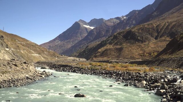 A cinematic wide establishing shot of a turquoise mountain river flowing through a massive, rocky Himalayan valley in Sissu, Lahaul, surrounded by barren peaks under a clear blue sky.