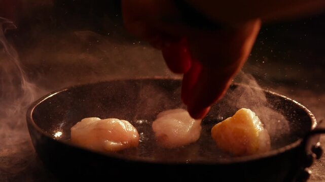 Hand seasoning scallops in a dark, smoky skillet during food preparation