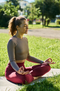 Vertical shot of young adult Caucasian woman meditating in lotus pose on green grass outdoors. Scene suitable for mindfulness apps, wellness marketing, and mental health campaigns