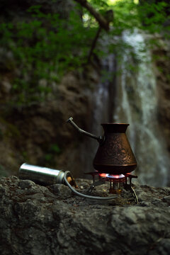 Small Turkish coffee pot on a portable camping stove with a gas canister, set against a scenic waterfall in a lush forest. Outdoor travel and coffee ritual concept.