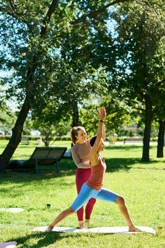 Vertical image of Caucasian instructor guiding African American woman practicing yoga warrior pose outdoors in park. Suitable for fitness branding and wellness app promotions