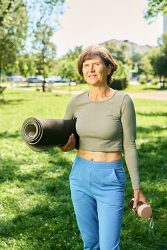 Portrait of middle aged Caucasian woman holding rolled yoga mat and water bottle in park. Image ideal for senior fitness campaigns and wellness marketing, vertical