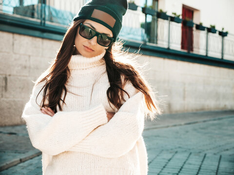 Portrait of beautiful smiling model. Female dressed in warm hipster white sweater and beanie. Trendy woman posing in the street at sunset in sunglasses