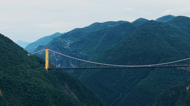 Grand aerial tracking right shot of a massive suspension bridge spanning across a deep mountain canyon under overcast sky.
