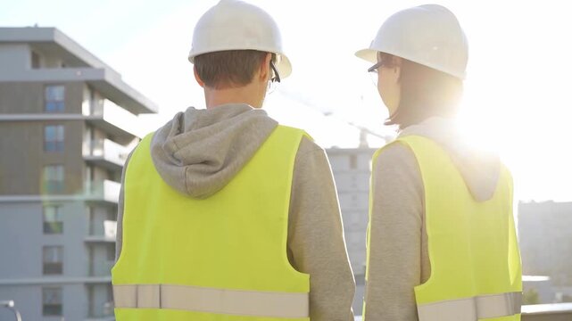 Man and woman architects or engineers wearing safety hard hats and vests holding blueprint and discussing something on construction site at sunrise, back view. Architecture and engineering