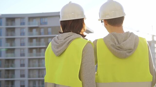 Man and woman architects or engineers wearing safety hard hats and vests holding blueprint and discussing something on construction site at sunrise, back view. Architecture and engineering