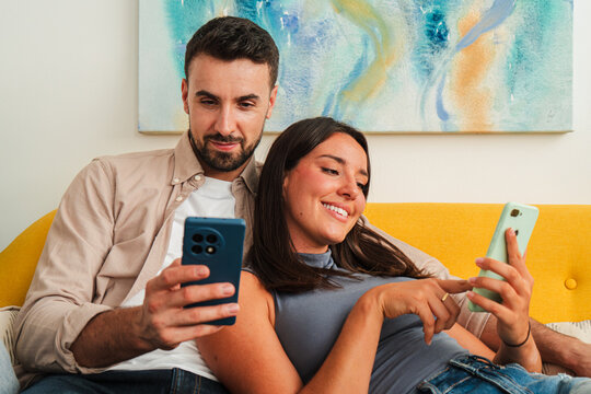 Two people sit close on a yellow sofa, scrolling and exchanging smiles while a pair of mobile phones shows content for both. Intimate, candid moment of shared digital life and joyful interaction.