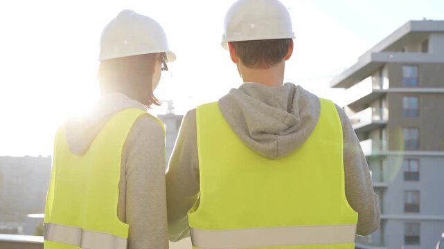 Man and woman architects or engineers wearing safety hard hats and vests holding blueprint and discussing something on construction site at sunrise, back view. Architecture and engineering